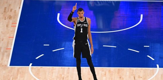 Victor Wembanyama, con la camiseta negra de los Spurs, levanta el dedo índice de pie en la zona de la pista