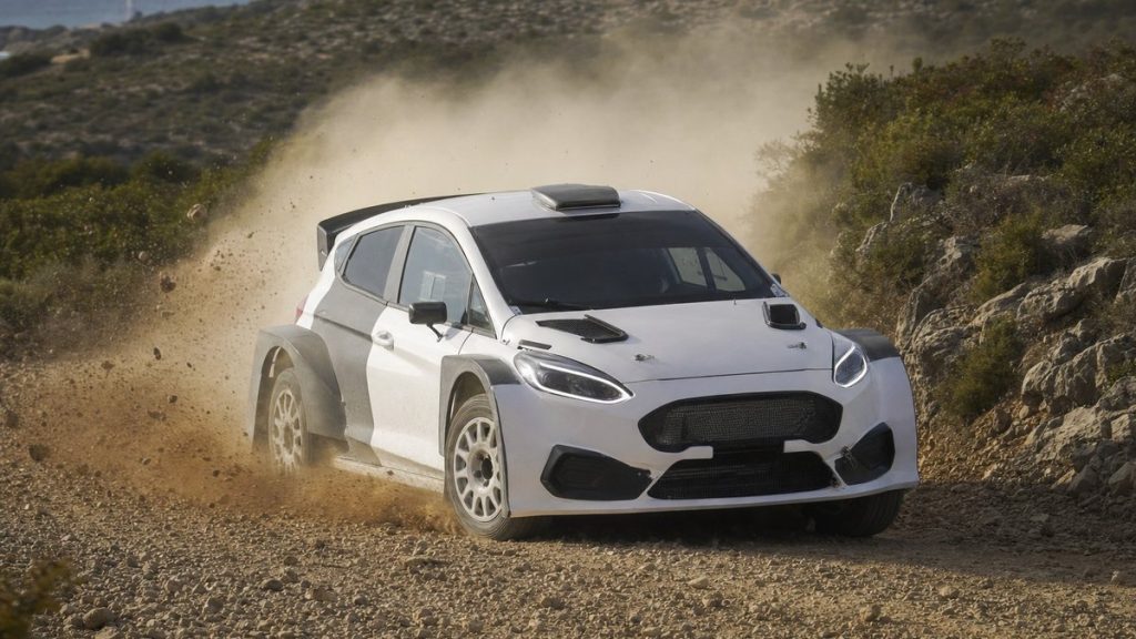 Un coche de rally blanco levanta polvo en un tramo de tierra entre matorrales y rocas