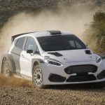 Un coche de rally blanco levanta polvo en un tramo de tierra entre matorrales y rocas