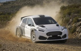 Un coche de rally blanco levanta polvo en un tramo de tierra entre matorrales y rocas