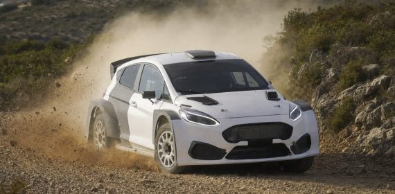 Un coche de rally blanco levanta polvo en un tramo de tierra entre matorrales y rocas