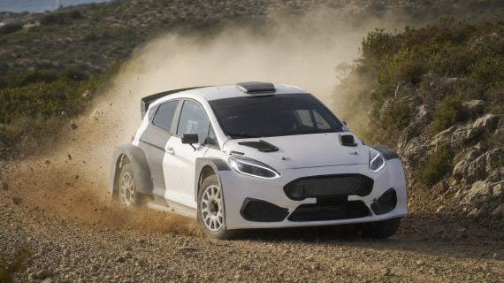 Un coche de rally blanco levanta polvo en un tramo de tierra entre matorrales y rocas