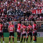 Varios jugadores del Athletic Club sobre el césped con la grada de los Bukaneros de fondo y un cartel de 'Presa, vete ya'.