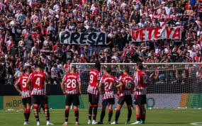 Varios jugadores del Athletic Club sobre el césped con la grada de los Bukaneros de fondo y un cartel de 'Presa, vete ya'.