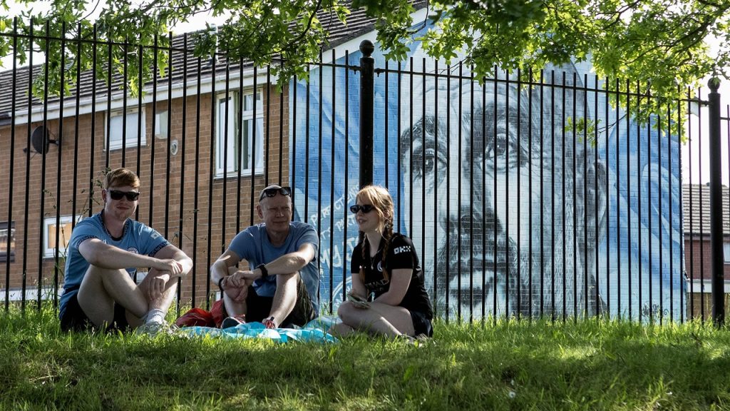 Aficionados del Manchester City sentados delante de un mural de Pep Guardiola durante la celebración del título de Premier League 2025/26.