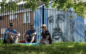 Aficionados del Manchester City sentados delante de un mural de Pep Guardiola durante la celebración del título de Premier League 2025/26.