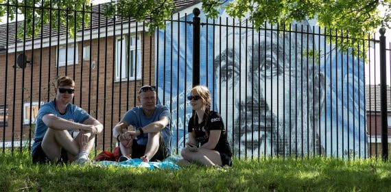 Aficionados del Manchester City sentados delante de un mural de Pep Guardiola durante la celebración del título de Premier League 2025/26.
